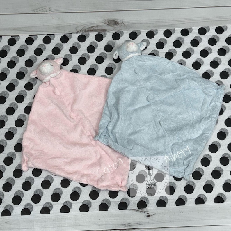 May include: Two plush baby blankets with lamb head attachments. One blanket is pink and the other is light blue. Both blankets have the name of the baby embroidered on them.