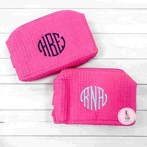 May include: Two hot pink cosmetic bags with a waffle texture. Each bag has a monogram in a contrasting color. One has a navy blue monogram, and the other has a light blue monogram. The bags are on a white wooden surface.