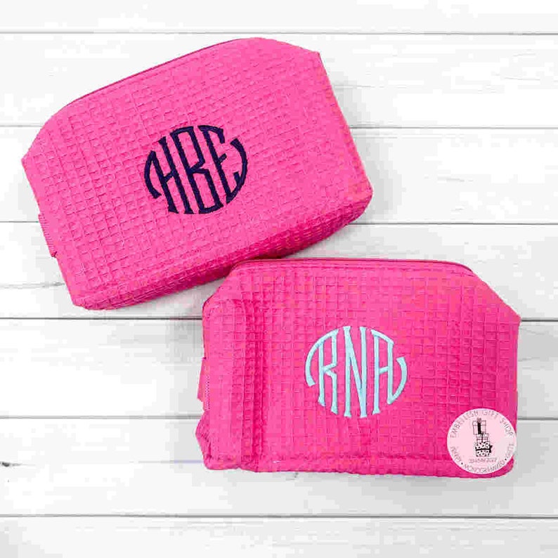 May include: Two hot pink cosmetic bags with a waffle texture. Each bag has a monogram in a contrasting color. One has a navy blue monogram, and the other has a light blue monogram. The bags are on a white wooden surface.