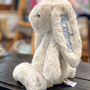 May include: A beige plush bunny with long floppy ears. The bunny is sitting on a wooden surface. The bunny's ear is embroidered with the name "Nicodemus" in blue thread.
