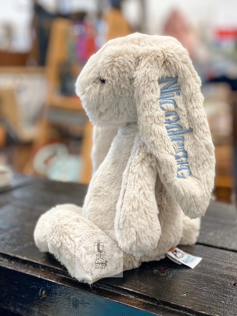 May include: A beige plush bunny with long floppy ears. The bunny is sitting on a wooden surface. The bunny's ear is embroidered with the name "Nicodemus" in blue thread.
