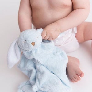以下が含まれることがあります： 赤ちゃんが青いぬいぐるみウサギのセキュリティブランケットを持っています。ブランケットは白い顔に茶色の目と茶色の鼻をしています。