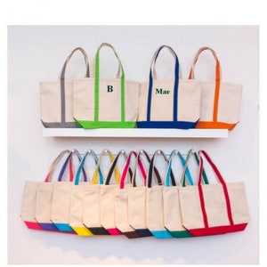 May include: A display of 17 canvas tote bags with colorful trim. The bags are arranged in two rows on a white shelf. The top row features three bags with personalized names, "B" and "Mae". The bottom row features 14 bags with a variety of colored trim.