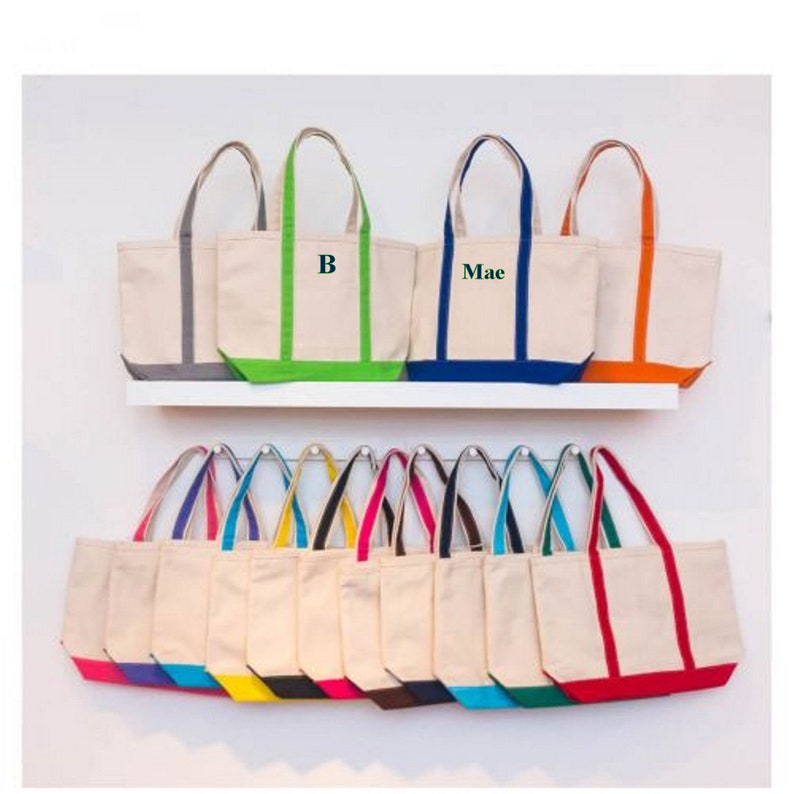 May include: A display of 17 canvas tote bags with colorful trim. The bags are arranged in two rows on a white shelf. The top row features three bags with personalized names, "B" and "Mae". The bottom row features 14 bags with a variety of colored trim.
