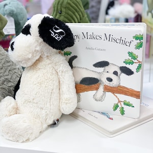 May include: A white plush dog toy with black ears and a black patch on its back, sitting next to a children's book titled "Puppy Makes Mischief" by Amelia Gatacre. The book features a black and white dog on a tree branch.