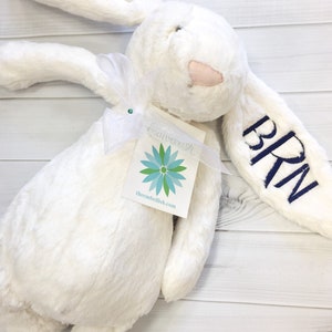 May include: White plush bunny with a blue embroidered name "BRN" on one ear. The bunny is holding a white tag with a blue flower design and the text "thembellish.com".