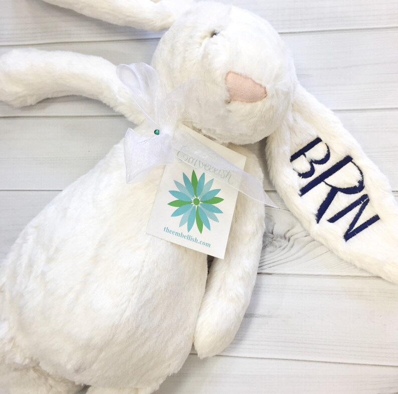 May include: White plush bunny with a blue embroidered name "BRN" on one ear. The bunny is holding a white tag with a blue flower design and the text "thembellish.com".