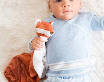May include: A baby wearing a light blue wrap-style top and striped pants, holding a plush fox toy. The fox toy is orange, white, and brown. The baby is lying on a white, fluffy surface.