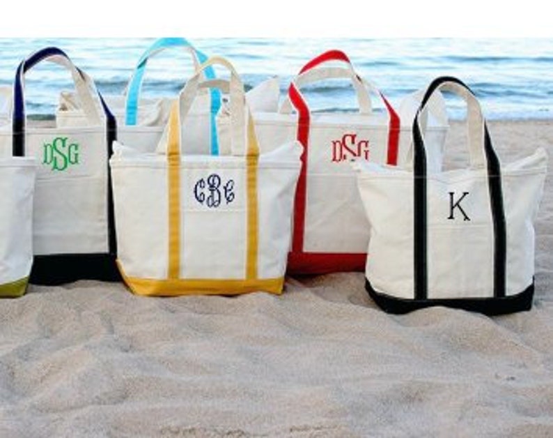 May include: A collection of six canvas tote bags with different colored trim and personalized initials. The bags are on a sandy beach.