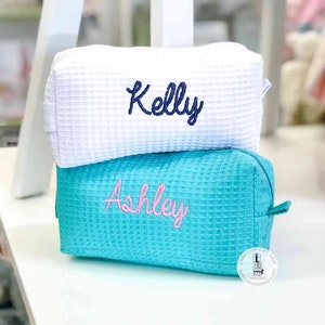 May include: Two white waffle-textured zippered pouches with embroidered names. The top pouch is white with the name "Kelly" embroidered in navy blue. The bottom pouch is turquoise with the name "Ashley" embroidered in pink.