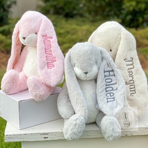 May include: Three plush bunny toys with long floppy ears. The bunnies are pink, gray, and cream colored. Each bunny has a name embroidered on one of its ears: Samantha, Holden, and Morgan.