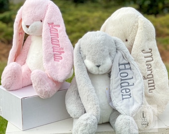 May include: Three plush bunny toys with long floppy ears. The bunnies are pink, gray, and cream colored. Each bunny has a name embroidered on one of its ears: Samantha, Holden, and Morgan.