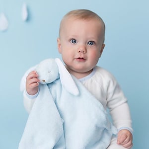May include: A baby is holding a blue and white bunny security blanket. The blanket has a soft, plush texture and a cute bunny face.