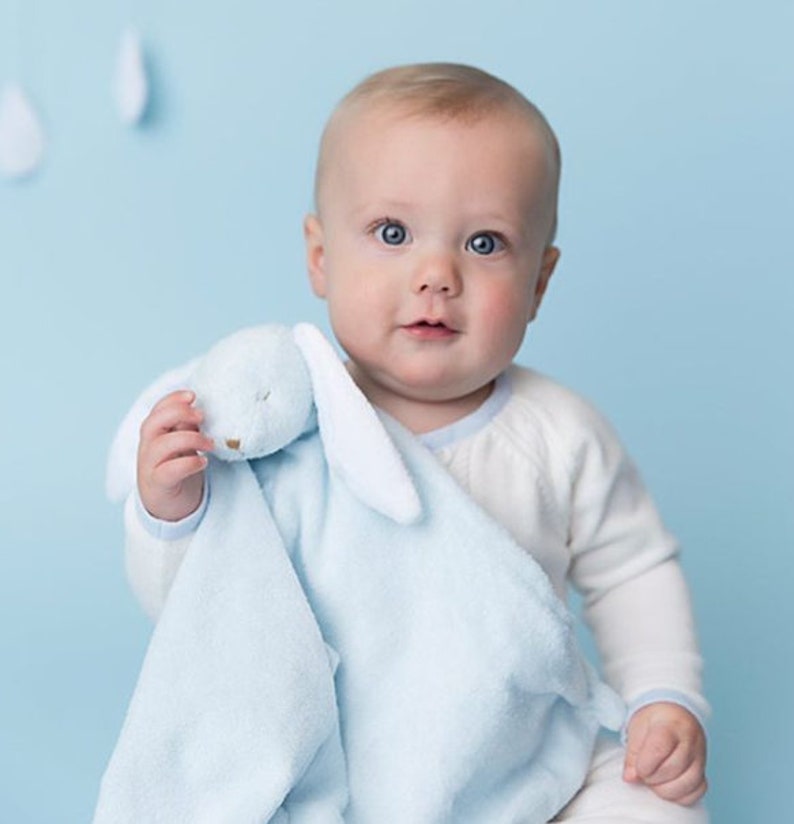 May include: A baby is holding a blue and white bunny security blanket. The blanket has a soft, plush texture and a cute bunny face.