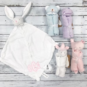 May include: A set of five plush baby toys, including a white bunny, a blue bear, a purple hippopotamus, a pink pig, and a white unicorn. Each toy is wrapped in a soft blanket and features embroidered details. The bunny blanket has the monogram "BGG" embroidered on it.