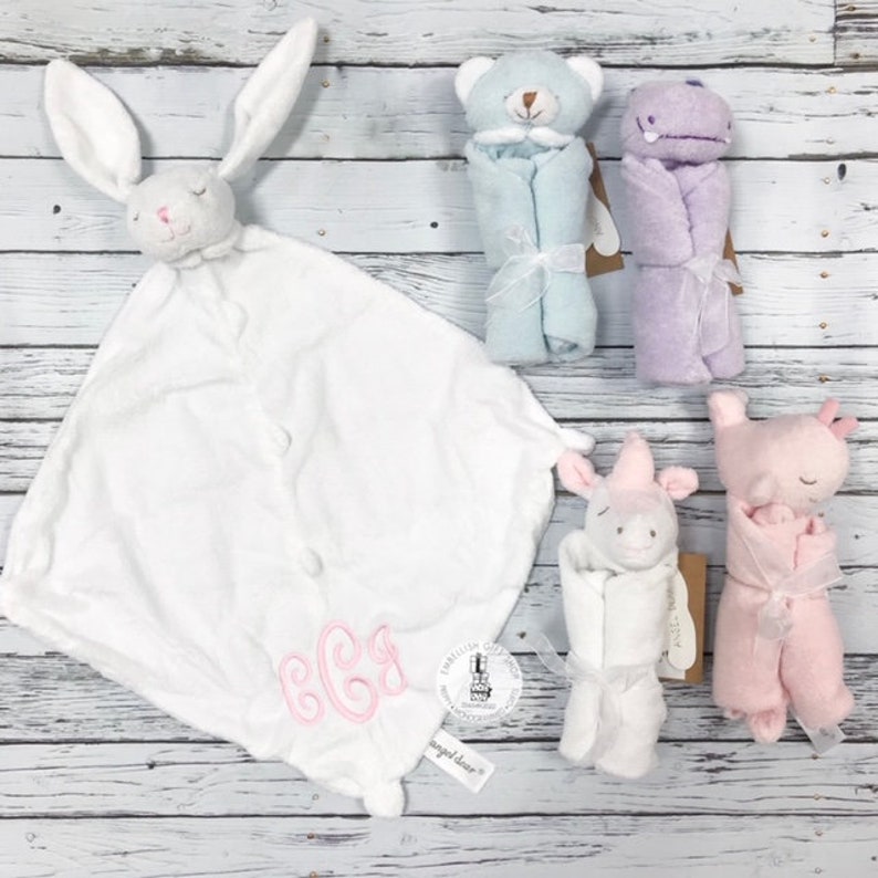 May include: A set of five plush baby toys, including a white bunny, a blue bear, a purple hippopotamus, a pink pig, and a white unicorn. Each toy is wrapped in a soft blanket and features embroidered details. The bunny blanket has the monogram "BGG" embroidered on it.