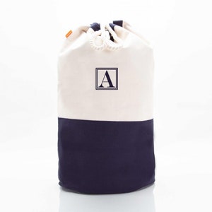 May include: A white canvas drawstring bag with a navy blue bottom. The bag has a rope drawstring and a navy blue square with the letter "A" in white.