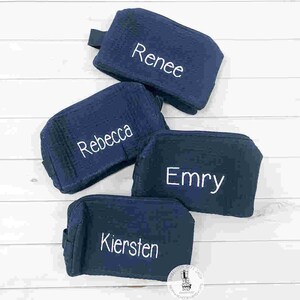 May include: Four navy blue cosmetic bags with white embroidered names: Renee, Rebecca, Emry, and Kiersten. Each bag has a small handle and a zipper closure. The bags are arranged on a white wooden surface.