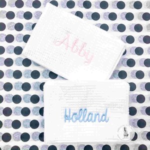 May include: Two white, textured rectangular items with embroidered names. One has "Abby" in pink script, the other has "Holland" in blue script. The background is a pattern of black and gray polka dots.