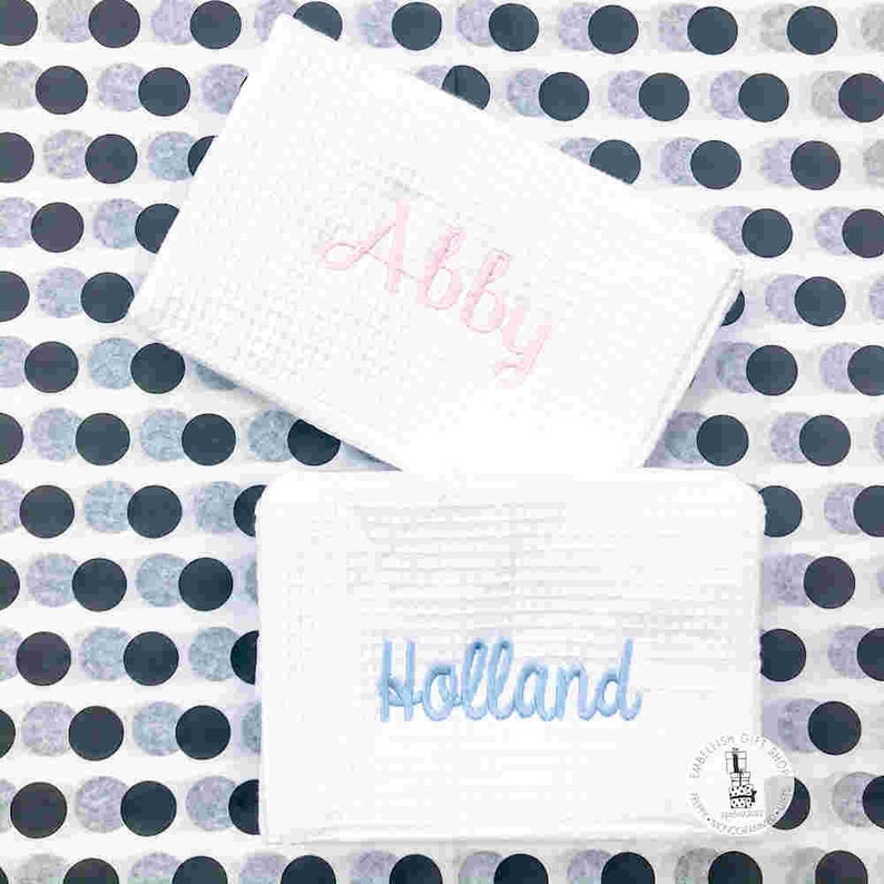 May include: Two white, textured rectangular items with embroidered names. One has "Abby" in pink script, the other has "Holland" in blue script. The background is a pattern of black and gray polka dots.