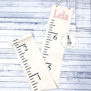 May include: A white canvas growth chart with black markings and a pink embroidered name "Lela". The chart is rolled up and has a black and white logo that says "Embellish Gift Shop Gifts 334.649.2022  Personalized Monogrammed".