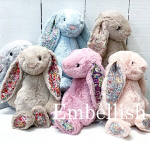May include: Five plush bunny toys in different colors, including light blue, beige, pink, and a darker shade of pink. Each bunny has floral patterned fabric on the inside of its ears and on the bottom of its feet. The bunnies are sitting on a white surface. The word "Embellish" is printed in purple on the surface.
