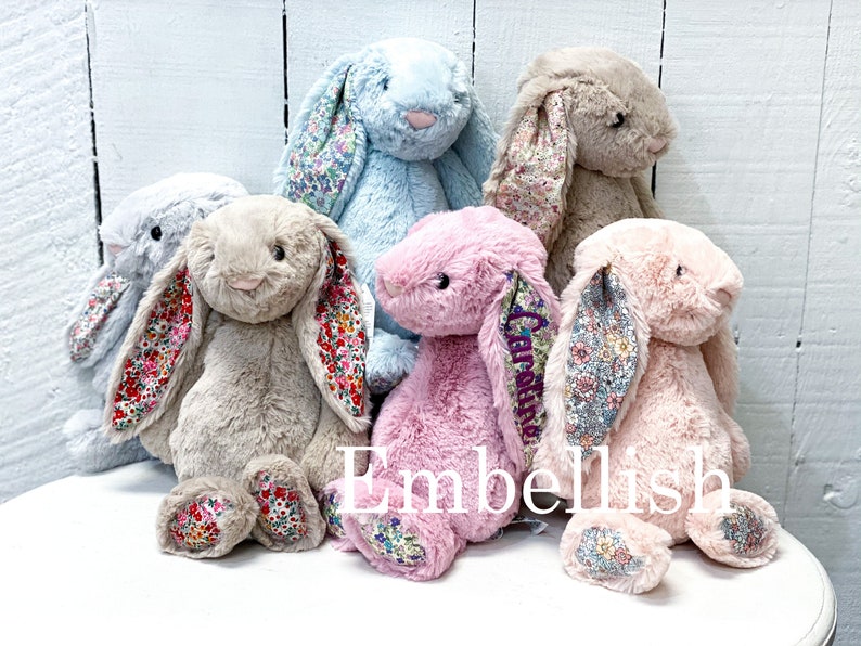May include: Five plush bunny toys in different colors, including light blue, beige, pink, and a darker shade of pink. Each bunny has floral patterned fabric on the inside of its ears and on the bottom of its feet. The bunnies are sitting on a white surface. The word "Embellish" is printed in purple on the surface.