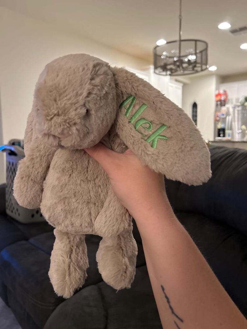 May include: A beige plush bunny with long ears. The bunny has a green embroidered name "Alek" on one ear.