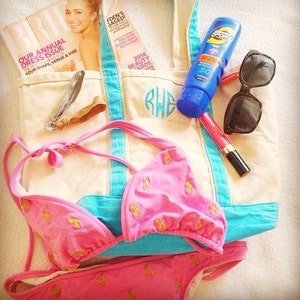 May include: A white canvas tote bag with blue trim and a monogrammed initial "RWE". The bag is filled with various items, including a pink bikini, a magazine, sunglasses, sunscreen, and lipstick.