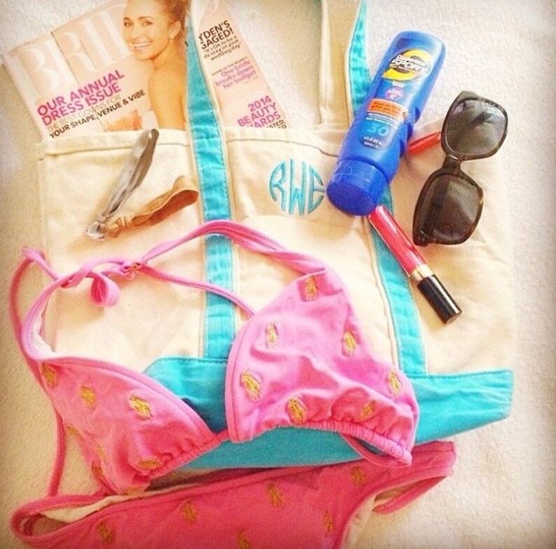 May include: A white canvas tote bag with blue trim and a monogrammed initial "RWE". The bag is filled with various items, including a pink bikini, a magazine, sunglasses, sunscreen, and lipstick.