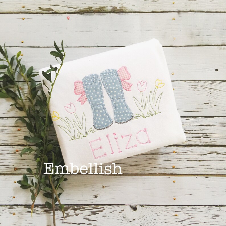 May include: A white embroidered t-shirt with a design of blue rain boots with pink bows, green grass, and yellow and pink tulips. The name "Eliza" is embroidered in pink below the design.