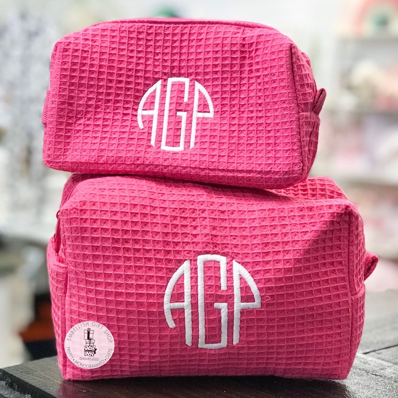 May include: Two stacked, hot pink cosmetic bags with a textured waffle weave pattern. Each bag features a white monogram with the letters "AGP". The bags have a rectangular shape with a zipper closure and small side pocket. The bags are for storing makeup and other beauty products.