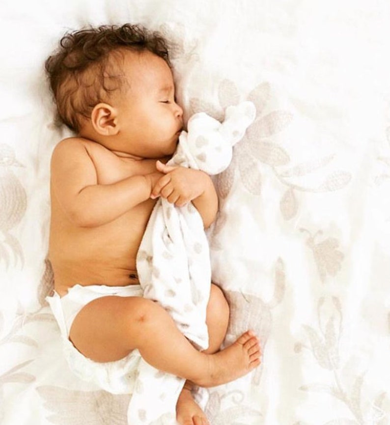 以下が含まれることがあります： 白い毛布の上で眠る赤ちゃん。白とグレーの斑点模様のぬいぐるみを抱えています。赤ちゃんは白いおむつをしています。