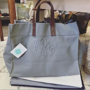 May include: A gray canvas tote bag with brown leather handles and a monogrammed "LSE" in white thread. The bag is sitting on a white surface.