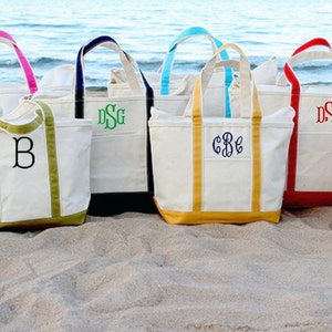 May include: Eight canvas tote bags with different colored trim and personalized monograms. The bags are lined up on a sandy beach.