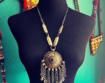 May include: A silver chain necklace with a large, ornate pendant. The pendant is circular and features a dark blue stone in the center, surrounded by smaller stones and intricate metalwork. The necklace is displayed on a black mannequin.