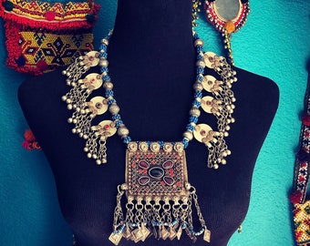 May include: A silver necklace with a large, square pendant featuring a dark blue stone surrounded by smaller red and white stones. The necklace is made of silver beads and has multiple silver tassels hanging from it.