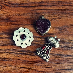 May include: Three silver pendants with intricate designs. One pendant features a red gemstone, another has an inscription, and the third has a cylindrical shape with dangling beads.