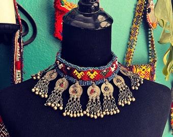 May include: A colorful beaded necklace with silver pendants and tassels. The necklace is on a black mannequin and has a red, blue, yellow, and green color scheme.