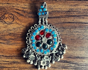 May include: A silver pendant with a round blue, green, and red gemstone design. The pendant has a decorative silver chain with small flower-shaped charms.
