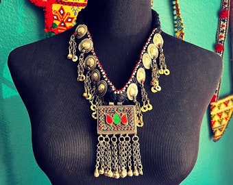 May include: A silver and black necklace with a rectangular pendant featuring red, green, and blue stones. The pendant is surrounded by a chain with multiple silver beads and charms.