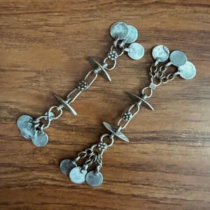 May include: A pair of silver-toned earrings with a vintage aesthetic. Each earring features a series of small, round, dangling charms and decorative chain links. The earrings have a detailed, handcrafted appearance, with a rustic finish.