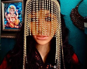 May include: A gold-toned rhinestone headpiece with a fringe of dangling rhinestones. The headpiece is worn by a person with long dark hair.