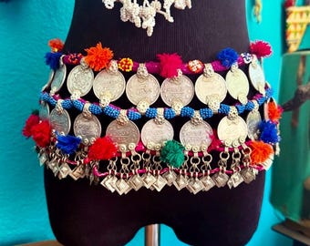 May include: A decorative waist belt featuring multiple rows of silver-toned coins, vibrant blue beaded strands, and small colorful yarn pom-poms in red, orange, pink, and green. The belt is constructed with a magenta fabric base and dangling charms.