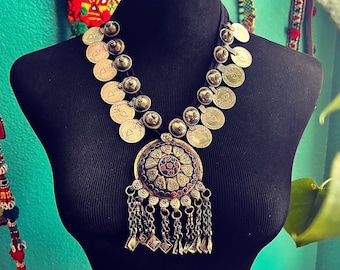 May include: A silver necklace with a large, round pendant featuring a colorful geometric design. The pendant is surrounded by smaller silver circles and hangs from a chain of silver circles.