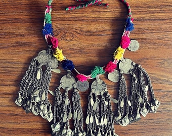 May include: A colourful beaded necklace with silver charms and tassels. The necklace is made with a variety of colours, including red, green, yellow, and blue. The charms are round and have a slightly textured surface. The tassels are long and flowy.