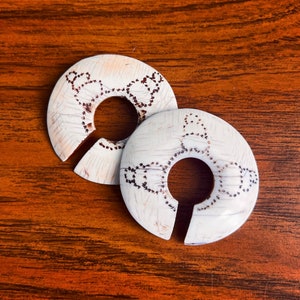 May include: Two off-white, circular wine bottle stoppers with a decorative, dotted design. Each stopper has a slot for the bottle neck. The stoppers are on a wooden surface.