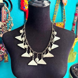 May include: Silver-toned necklace with triangular and heart-shaped pendants. The necklace is displayed on a black mannequin. The background features colorful textiles and a turquoise wall.