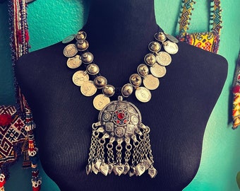 May include: A silver necklace with a large pendant featuring a red gemstone surrounded by smaller silver pieces. The necklace is made of a series of silver coins with intricate designs.