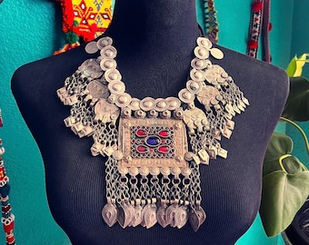 May include: A silver necklace with a large, rectangular pendant featuring colorful stones. The pendant is surrounded by smaller silver circles and chains. The necklace is displayed on a black mannequin.